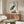 May include: Japanese woodblock print of grey heron with orange beak standing among marsh grasses, featuring traditional calligraphy, displayed in thin brown frame above mid-century walnut credenza. White curved chair and glass-top coffee table visible in minimalist room with cream walls and natural lighting.