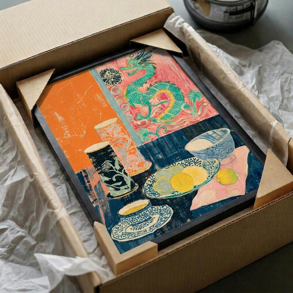 May include: Framed art print in black frame being carefully unpacked from cardboard shipping box with protective tissue paper. The colorful artwork features a vibrant still life with Asian-inspired elements including a green dragon motif, orange and coral backgrounds, blue patterned ceramics, and yellow eggs or fruit. The piece shows bold colors and appears to be in a vintage or woodblock print style.
