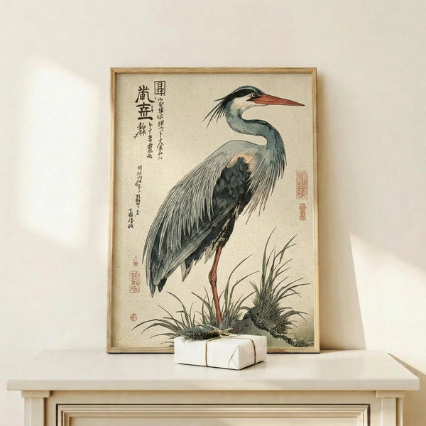 May include: Japanese woodblock print of a grey heron with long orange beak and legs standing among marsh grasses, featuring traditional calligraphy and red stamps, displayed in light wooden frame on pale wood console table next to small white wrapped gift box against cream wall