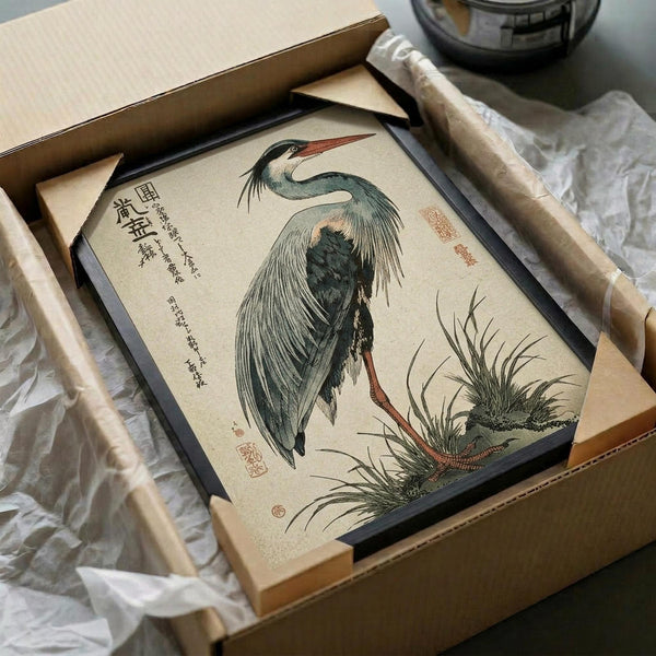 May include: Framed vintage Japanese woodblock print of a grey heron with long neck and flowing feathers standing among reeds, featuring traditional Asian calligraphy and red seal stamps. The artwork is displayed in a black frame within an open cardboard shipping box with protective tissue paper, surrounded by wooden frame corners and packaging materials on a white surface.