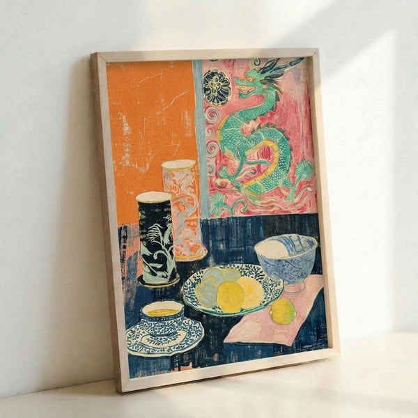 Framed art print leaning against white wall showing a colorful still life scene with Asian-inspired decorative elements. The composition features ceramic vessels, bowls with fruit, and a teacup on a dark blue table surface. An ornate green dragon motif decorates the pink and coral background wall. The artwork combines bold orange, pink, blue and green colors in a modern artistic style with decorative patterns throughout.