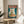 May include: Framed poster print featuring a white cat with golden eyes eating ramen noodles from a blue bowl, with red Japanese characters above on teal background. Displayed on light wood round side table next to beige upholstered chair with throw pillow. Natural sunlight streams through window, casting shadows from nearby plant leaves onto white wall.