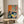 May include: Framed colorful still life artwork displayed on round wooden side table next to beige upholstered chair. Print features vibrant orange and pink background with green dragon motif, dark blue table surface holding yellow lemons on decorative plate, ornate teacup, ceramic vases, and small bowl. Natural lighting creates soft shadows across white wall and plant leaves visible at edge.