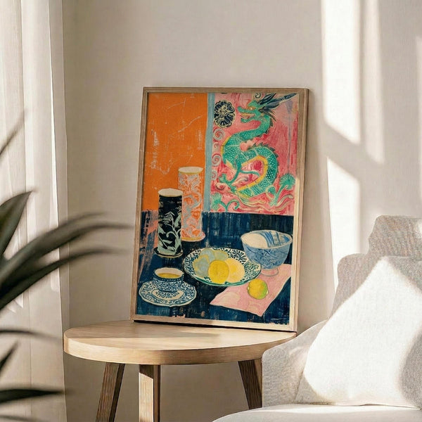May include: Framed colorful still life artwork displayed on round wooden side table next to beige upholstered chair. Print features vibrant orange and pink background with green dragon motif, dark blue table surface holding yellow lemons on decorative plate, ornate teacup, ceramic vases, and small bowl. Natural lighting creates soft shadows across white wall and plant leaves visible at edge.