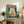 May include: Framed poster featuring a white cat with yellow eyes eating ramen noodles from a blue bowl, with red Japanese characters on teal background, displayed on rustic wooden sideboard alongside books, ceramic vase, and fern plant in cozy living room setting with beige armchair visible.