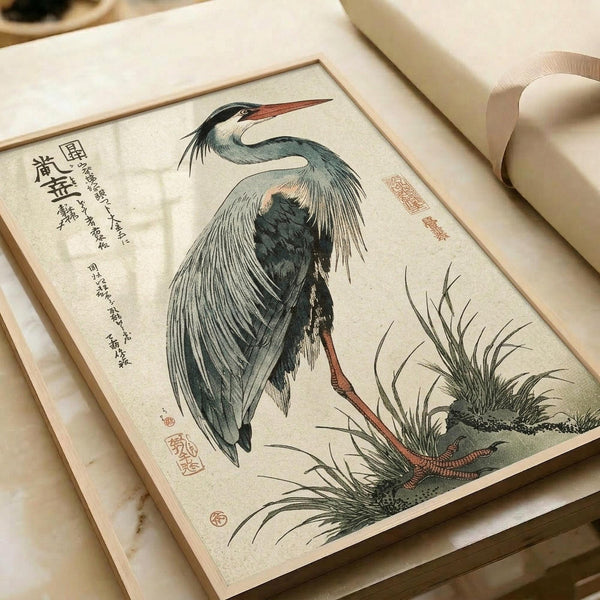 A vintage Japanese woodblock print depicting a grey heron with dark plumage and reddish-orange beak and legs, standing among marsh grasses. The artwork features traditional muted colors in blue-grey and green tones against a cream background, with Japanese calligraphy and red seal stamps. The print is displayed in an open book or portfolio with aged paper pages and a cream ribbon bookmark visible.