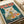 May include: Vintage-style poster print featuring a white cat with golden eyes eating ramen noodles with chopsticks from a blue bowl containing eggs, meat, and vegetables. Red Japanese characters appear at the top against a teal background. The artwork is displayed on white paper with rolled paper tubes visible in the background, showing the print's flat poster format.