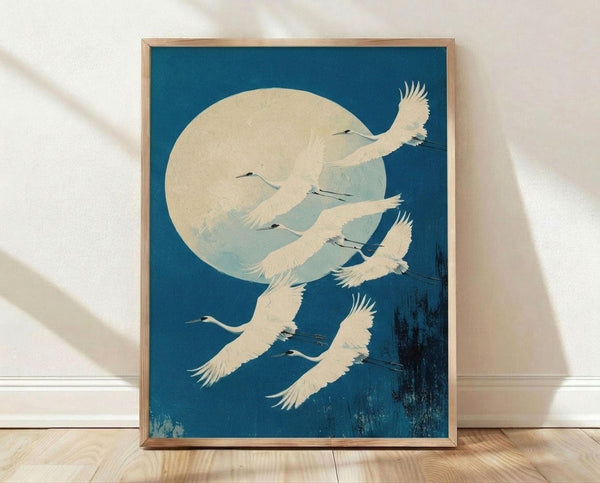 May include: Framed vintage Japanese woodblock print showing white cranes flying against deep blue sky with large pale moon. Art features delicate feather details and subtle brushwork textures. Natural wood frame sits on light wooden floor against white textured wall with soft shadows.