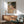 May include: Framed art print of an expressionist painting showing intertwined nude figures in muted earth tones with bold black outlines, displayed above a mid-century modern wooden credenza with lamp, next to a sage green upholstered chair in a sunlit room with white walls.