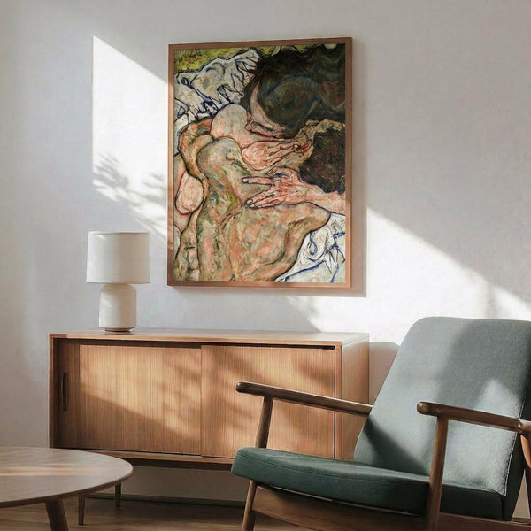 May include: Framed art print of an expressionist painting showing intertwined nude figures in muted earth tones with bold black outlines, displayed above a mid-century modern wooden credenza with lamp, next to a sage green upholstered chair in a sunlit room with white walls.
