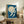 May include: Framed artwork displaying white cranes flying against a deep blue sky with large pale moon, positioned on rustic wooden sideboard alongside potted fern, stacked books, and ceramic vessels in neutral-toned living room setting