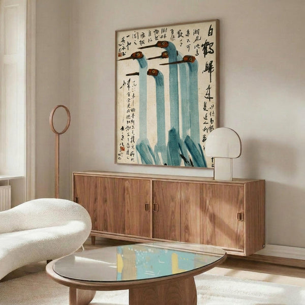 May include: Chinese-style artwork featuring five blue cranes with long necks and dark beaks against cream background with black calligraphy text, displayed in thin wood frame above walnut credenza. Modern living room setting with white curved chair, brass floor lamp, oval glass coffee table, and neutral walls.