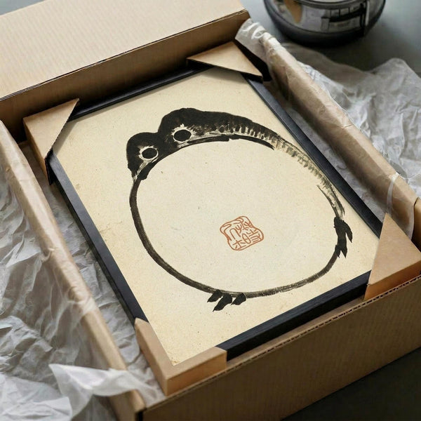A minimalist Japanese ink painting of a round frog viewed from above, painted in bold black brushstrokes on cream paper with a small red seal stamp. The artwork is displayed in a black frame inside an open brown cardboard box with white tissue paper, surrounded by wooden dowels and packaging materials on a white surface.