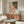May include: Expressionist artwork in warm earth tones depicting embracing figures, displayed above a mid-century walnut credenza in a modern living room. The painting features flowing, organic forms in pink, green, and cream hues with bold brushstrokes. White sculptural chair and oval glass coffee table complete the contemporary interior setting with neutral walls and natural lighting.