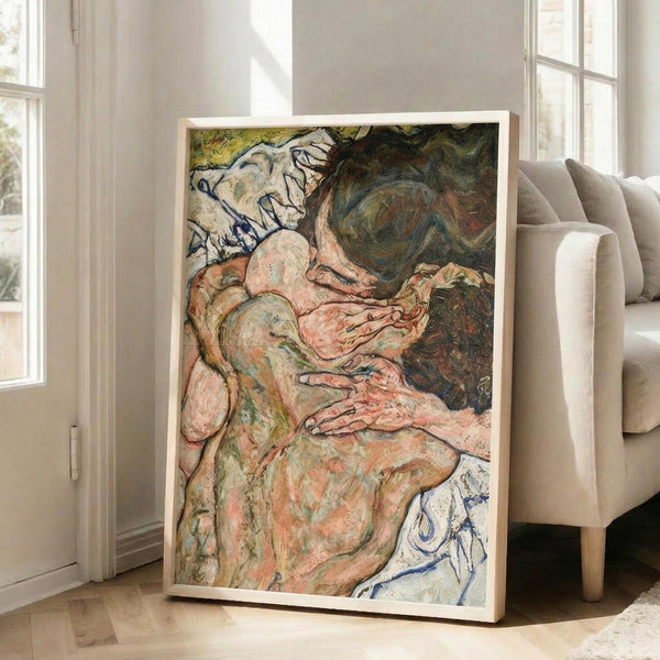 May include: Framed reproduction of an expressionist painting showing two embracing nude figures rendered in earthy tones of pink, green, and brown with bold black outlines. The artwork is displayed in a light wood frame leaning against a beige armchair in a bright living room with white walls and large windows.