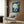 May include: Japanese-style art print showing white cranes flying against deep blue sky with large pale moon, displayed in light wood frame on textured brown wall panel above cream upholstered armchair with geometric patterned pillow, brass floor lamp and side table with open book nearby