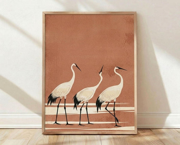 May include: Framed poster print featuring three white cranes with black wing tips and long dark beaks standing on pale horizontal stripes against a warm terracotta brown background. The artwork has a minimalist Japanese-inspired style with subtle texture. Wooden frame sits on light wood flooring against white fabric backdrop with soft natural lighting and shadows.