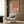 May include: Framed poster with three white cranes on terracotta orange background displayed on wood-paneled wall above beige upholstered armchair with geometric patterned pillow. Modern reading nook with brass floor lamp, round side table with open book, and sheer curtains. Natural lighting creates warm, minimalist interior setting.