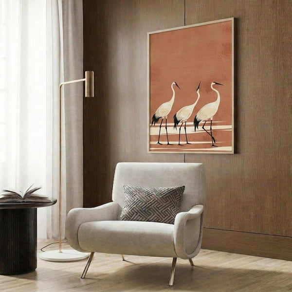 May include: Framed poster with three white cranes on terracotta orange background displayed on wood-paneled wall above beige upholstered armchair with geometric patterned pillow. Modern reading nook with brass floor lamp, round side table with open book, and sheer curtains. Natural lighting creates warm, minimalist interior setting.