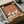 May include: Framed artwork featuring three elegant white cranes with black-tipped feathers and long curved necks, standing on geometric striped ground in warm terracotta and cream tones. The piece sits in a dark frame within brown cardboard packaging surrounded by white tissue paper, with wooden craft sticks visible nearby on a neutral surface.