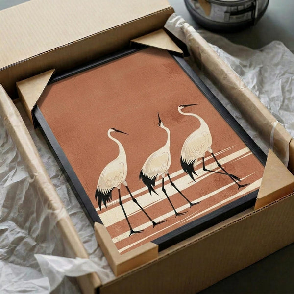 May include: Framed artwork featuring three elegant white cranes with black-tipped feathers and long curved necks, standing on geometric striped ground in warm terracotta and cream tones. The piece sits in a dark frame within brown cardboard packaging surrounded by white tissue paper, with wooden craft sticks visible nearby on a neutral surface.