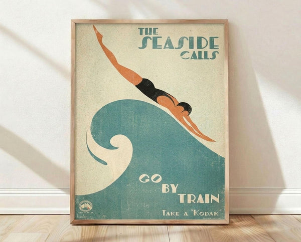 May include: Vintage-style travel poster in light wood frame leaning against white wall on wooden floor. Features art deco illustration of swimmer diving over stylized teal wave on cream background. Text reads "The Seaside Calls" and "Go By Train" with Kodak branding. Poster has weathered, aged appearance with muted teal and orange colors in retro tourism advertisement style.