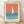 May include: Framed vintage-style poster print featuring minimalist sailboat illustration with white sail on coral orange sky and teal blue water, displayed in light wood frame against white wall on wooden floor with natural lighting and soft shadows
