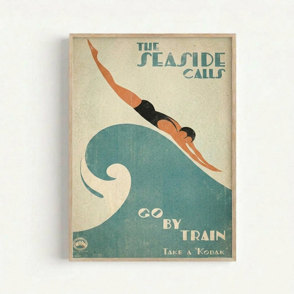 Vintage-style travel poster with art deco design featuring a swimmer diving over a large teal wave. Text reads "The Seaside Calls" at top and "Go By Train" and "Take a Kodak" below. Cream background with coral, black and teal colors. Displayed in light wood frame against white wall.