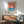 May include: Framed vintage-style poster featuring a sailboat on blue water against coral-orange sky, displayed above a mid-century modern wooden credenza with table lamp, next to a sage green upholstered armchair in a sunlit room with white walls