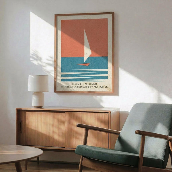 May include: Framed vintage-style poster featuring a sailboat on blue water against coral-orange sky, displayed above a mid-century modern wooden credenza with table lamp, next to a sage green upholstered armchair in a sunlit room with white walls