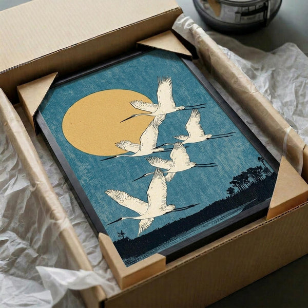 May include: Framed vintage Japanese woodblock print showing white cranes flying across blue sky with golden moon, dark silhouetted landscape below. Artwork displayed in black frame, partially unwrapped from brown shipping box with protective tissue paper on white surface.