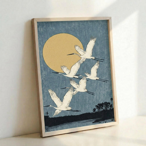 May include: A framed vintage Japanese woodblock print showing six white cranes flying diagonally across a blue sky with a large golden moon. The birds have detailed feathers and long necks, soaring above a dark silhouetted landscape with trees and water. The artwork is displayed in a light wood frame, leaning against a white wall on a white surface, showcasing the traditional ukiyo-e art style with muted blue, cream, and black tones.