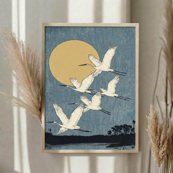 May include: Framed vintage Japanese woodblock print featuring five white cranes flying against a blue textured sky with large golden moon. Dark silhouetted landscape with trees and water below. Light wooden frame displayed on white wall beside dried pampas grass in neutral-toned room setting with natural lighting.