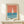 May include: Framed vintage-style poster featuring a minimalist sailboat design with coral orange sky, teal blue water, and white sail. The print shows "MADE IN USSR IMPREGNATED SAFETY MATCHES" text at bottom. Displayed on light wood surface with small wrapped gift beside frame. Natural lighting creates soft shadows on cream wall background.