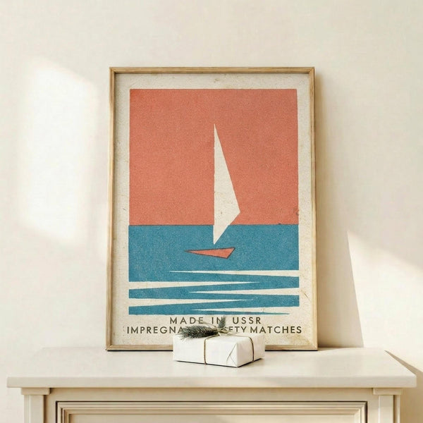 May include: Framed vintage-style poster featuring a minimalist sailboat design with coral orange sky, teal blue water, and white sail. The print shows "MADE IN USSR IMPREGNATED SAFETY MATCHES" text at bottom. Displayed on light wood surface with small wrapped gift beside frame. Natural lighting creates soft shadows on cream wall background.