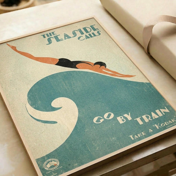 May include: Vintage-style travel poster print displayed on white surface with rolled paper tubes nearby. Features art deco illustration of swimmer diving over stylized teal wave with cream background. Text reads "THE SEASIDE CALLS" at top and "GO BY TRAIN" with "TAKE A KODAK" below. Weathered, aged appearance with muted orange, teal, and cream color palette in classic 1930s advertising poster style.