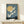 May include: Framed vintage Japanese art print displaying white cranes in flight against dusty blue sky with golden moon, dark silhouetted landscape below. Artwork has textured appearance in thin wooden frame, displayed on light wood surface alongside small wrapped gift box with natural twine and dried plant sprig.