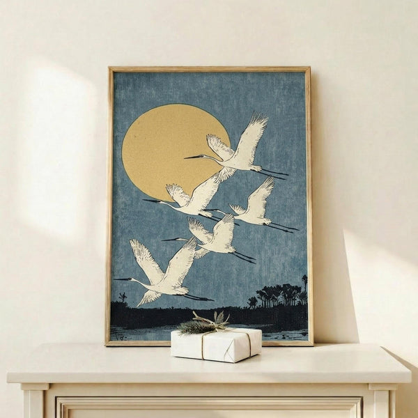 May include: Framed vintage Japanese art print displaying white cranes in flight against dusty blue sky with golden moon, dark silhouetted landscape below. Artwork has textured appearance in thin wooden frame, displayed on light wood surface alongside small wrapped gift box with natural twine and dried plant sprig.