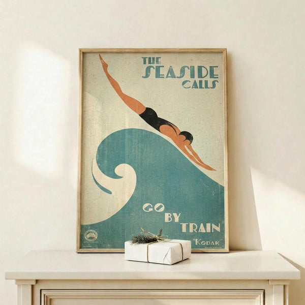 May include: Vintage travel poster in wooden frame featuring art deco style illustration of swimmer diving into large teal wave. Text reads "The Seaside Calls" and "Go By Train" with Kodak branding. Poster displayed on light wood shelf with small wrapped gift package, against white wall with natural lighting creating soft shadows.