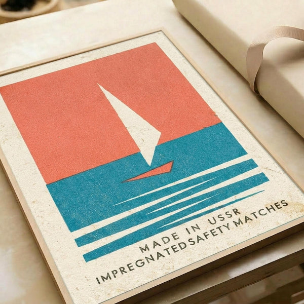 May include: Vintage-style poster print featuring a geometric sailboat design with white sail against coral red sky and teal blue water with horizontal wave lines. Text reads "MADE IN USSR IMPREGNATED SAFETY MATCHES" below the nautical illustration. Print shown in natural lighting on cream paper with visible texture, displayed alongside rolled papers and ribbon on wooden surface.