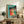 May include: Framed vintage-style poster featuring five dancing women in colorful dresses (red, orange, yellow, green, blue) with "Dance More" text on turquoise background, displayed on rustic wooden sideboard alongside books, ceramic vases, fern plant, and leather furniture in cozy living room setting with neutral walls.
