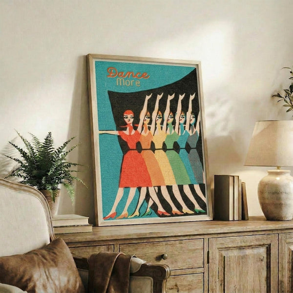 May include: Framed vintage-style poster featuring five dancing women in colorful dresses (red, orange, yellow, green, blue) with "Dance More" text on turquoise background, displayed on rustic wooden sideboard alongside books, ceramic vases, fern plant, and leather furniture in cozy living room setting with neutral walls.