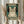 May include: Person holding framed poster featuring tabby cat wearing black headphones on teal vintage-style background with cream text. Wooden frame, neutral room setting with dried pampas grass visible. Hands wearing cream ruffled sleeves hold rectangular wall art print.