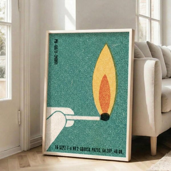 May include: Framed vintage-style poster with textured teal background featuring a white hand holding a lit match with yellow and orange flame. The minimalist design includes Polish text at bottom and appears to be a reproduction of a 1960s matchbox label. The wooden-framed print leans against a white wall beside a beige upholstered sofa in a bright, modern living room with large windows.