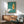 May include: Vintage poster with turquoise background featuring stylized white matchstick and orange flame, displayed in wooden frame above mid-century modern walnut credenza. Green upholstered armchair with wooden arms sits beside small round table with ceramic lamp. Bright natural light creates geometric shadows on white wall in minimalist living room setting.