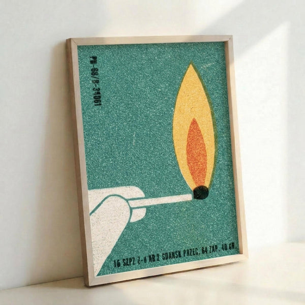 Vintage-style poster with textured teal background featuring a white hand holding a lit match with yellow and orange flame. Text appears in Polish at top and bottom. The artwork is displayed in a light wood frame leaning against a white wall, showing the grainy, matchbox label aesthetic with muted colors and retro typography.