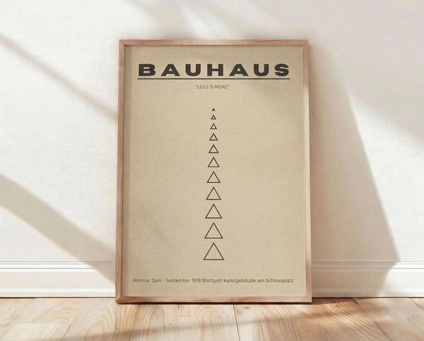 May include: Framed beige poster with bold black "BAUHAUS" text and "LESS IS MORE" quote, featuring a vertical row of minimalist triangular shapes in varying sizes. Natural wood frame displayed against white textured wall on light wooden floor with soft natural lighting creating gentle shadows.