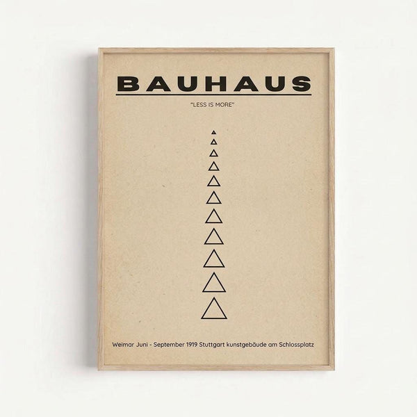 A minimalist beige poster in a light wood frame displays "BAUHAUS" in bold black letters at the top, followed by "LESS IS MORE" in smaller text. Below, eleven black triangular outlines of varying sizes are arranged vertically in a column. At the bottom, small black text reads "Weimar Juni - September 1919 Stuttgart kunstgebäude am Schlossplatz". The framed artwork hangs on a white wall.