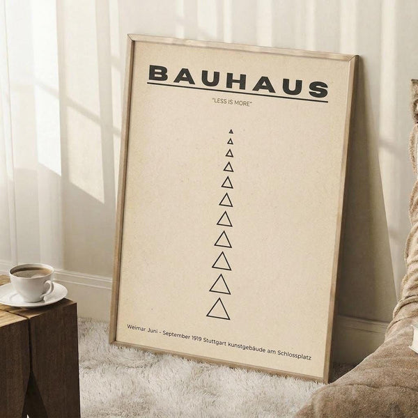 May include: Bauhaus-style minimalist poster in light wood frame leaning against white wainscoted wall. Cream background with bold black text reading "BAUHAUS" and "LESS IS MORE" above vertical row of geometric triangles. German exhibition text at bottom. Shown with coffee cup on wooden block and cozy beige throw blanket on white textured rug.