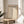 May include: Minimalist Bauhaus poster with cream background and black geometric triangles in vertical arrangement, displayed in thin wooden frame on round wooden side table next to beige upholstered chair. Soft natural lighting creates shadows across white walls, with green plant leaves visible in corner of modern neutral-toned room setting.